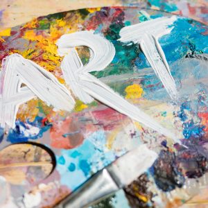 Word art painted with white gouache on colorful palette with paintbrush near by